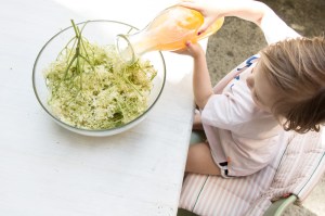 Kind gießt Apfelsaft über Holunderblüten, sitzt dabei auf Tripp Trapp in den neuen Farbe 2017 Moss Green
