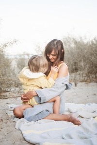 Mutter und Kind innig am Strand. Leinentuch udn Bademantel von Kaisu Mari, Badeanzug One Shoulder von Mymarinin, Kinderbademantel von Trixie baby über Nickis.com