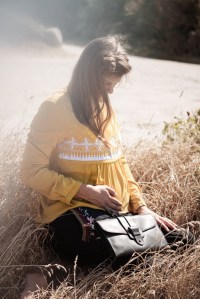 Schwangere Frau trägt Langarmshirt mit Stillfunktion in Senfgelb, dazu Umstandshose von mara Mea udn Clutch von Planet Hiitu